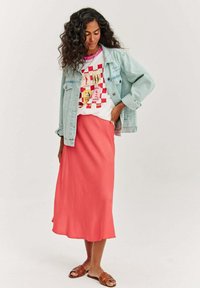 Dżinsowa kurtka, biała koszulka z grafiką w czerwone akcenty, zwiewna koralowa spódnica oraz brązowe sandały na paski, tworzące luźny, warstwowy wygląd.