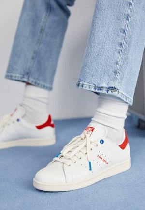 Baskets blanches avec accents rouges et œillets bleus portées avec des chaussettes blanches et un jean en denim bleu clair, debout sur un sol recouvert d'un tapis bleu.