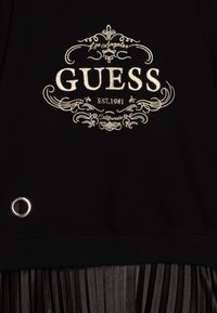 Schwarzer Pullover mit goldbesticktem "GUESS"-Logo, ornamentalem Text und kreisförmigem silbernem Ösen-Detail am Saum.