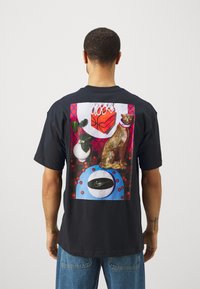 Camiseta negra de manga corta con un gráfico colorido en la parte posterior, que representa un gato dorado, balones de baloncesto y formas estilizadas.