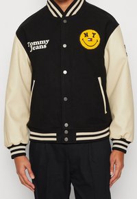Černá vlněná varsity bunda s krémovými koženými rukávy, pruhovanými manžetami a límcem. Obsahuje vyšitý žlutý smajlík a logo "Tommy Jeans".