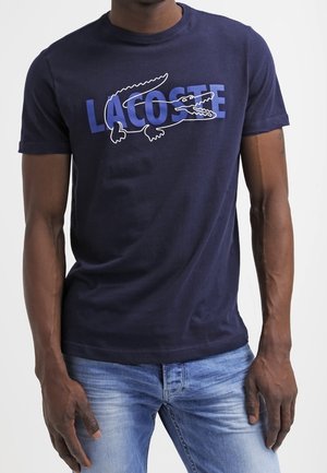 Mężczyzna noszący granatowy t-shirt z niebieskim napisem „LACOSTE” i białym obramowaniem logo krokodyla, w połączeniu z jasnoniebieskimi jeansami.