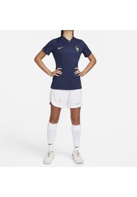 Maillot de soccer bleu marine avec un col et des accents dorés, associé à un short blanc avec un logo doré, des chaussettes blanches et des chaussures blanches Nike.