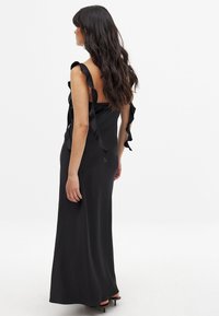 Vestido maxi negro sin mangas con tirantes con volantes, textura suave y un corsé ajustado; la vista trasera muestra una silueta de longitud completa y detalles delicados.