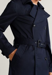 Mężczyzna ubrany w ciemnogranatowy płaszcz trencz z paskiem, zapinany na podwójne guziki, z ręką w kieszeni, pokazany od ramion do połowy ud.