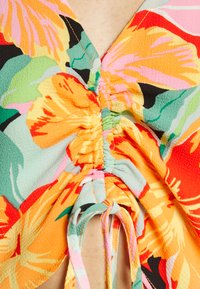 Top floral com cores vibrantes como laranja, verde e rosa. Apresenta um tecido texturizado, detalhe drapeado na gola e laços.