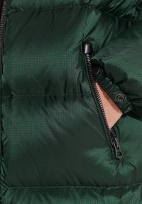 Sezione di giacca a vento verde scuro con zip, tasca con cerniera, tessuto testurizzato e polsini elasticizzati sicuri per una vestibilità aderente.