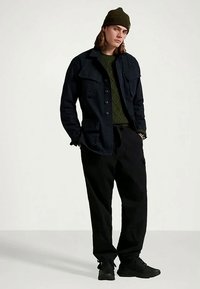 Νεαρός άνθρωπος που φοράει ένα σκούρο καπέλο, μαύρο μπουφάν πάνω από πράσινο πουλόβερ, μαύρο παντελόνι και μαύρα παπούτσια, στέκεται με τα χέρια στις τσέπες.