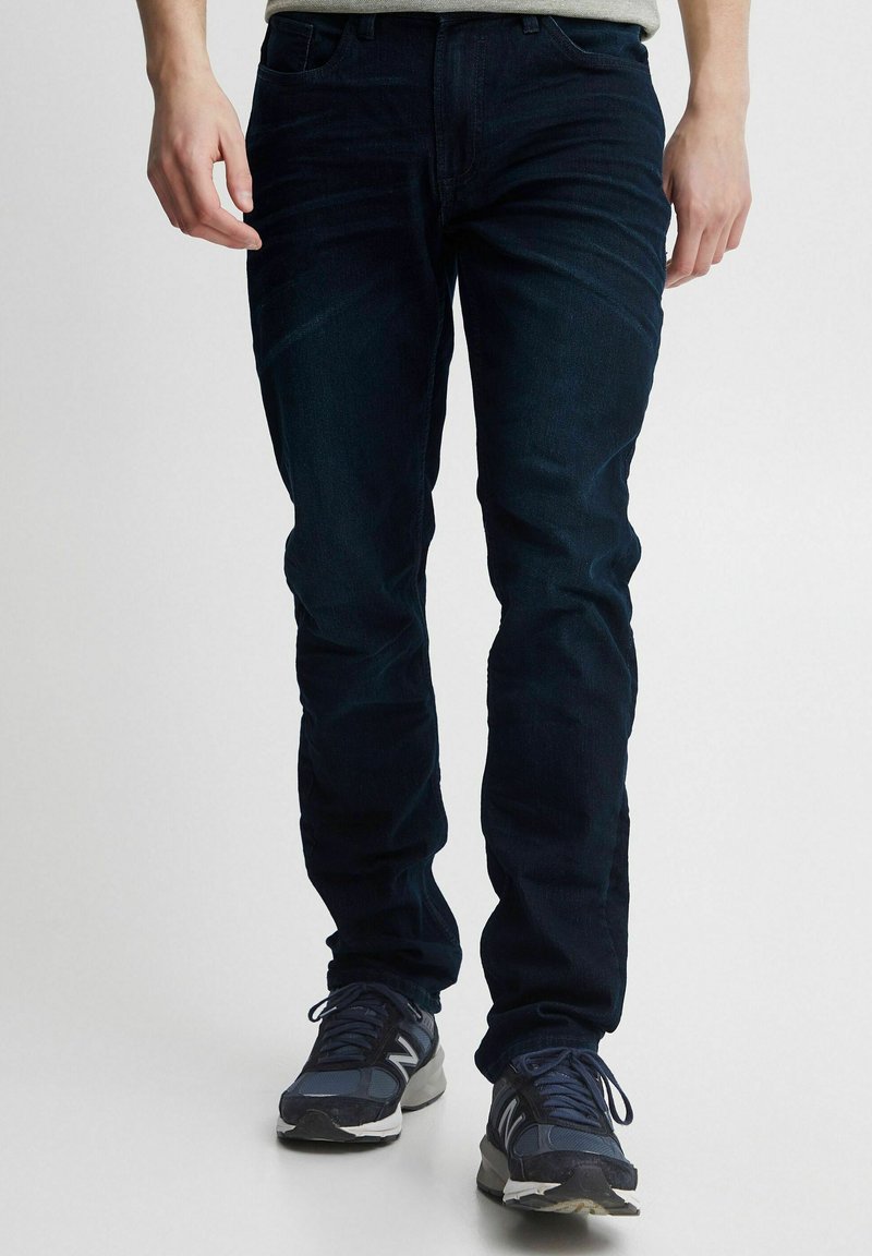 Tumedat sinised denim püksid, millel on kitsas lõige, viis taskut ja õrn fade efekt. Kantud tumesiniste spordijalatsitega, et saavutada juhuslik välimus.