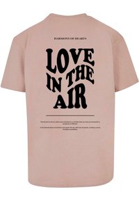 Camiseta de algodón rosa con texto negro en la parte posterior: "AMOR EN EL AIRE" con una fuente decorativa y una cita debajo. Mangas cortas, corte relajado.