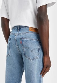 Personne portant un jean Levi's bleu clair et un t-shirt blanc, vue de dos, avec une bague à la main droite.