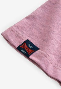Rosa bomull t-shirt tyg med en slät textur, med en marinblå etikett med röd text. Etiketten läser "Superdry" och "Trade Mark."