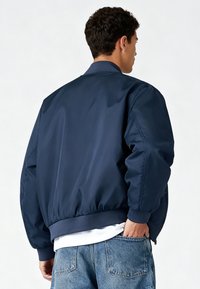 Veste bomber marine avec une finition lisse et brillante, poignets et ourlet côtelés, silhouette décontractée, associée à un jean bleu clair.