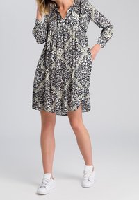 Robe à motifs noir et blanc avec des manches longues, présentant une coupe ample, un décolleté en V, et agrémentée de dessins géométriques verts, associée à des baskets blanches.