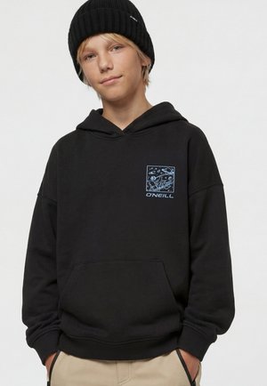 Junge Person trägt eine schwarze Mütze und einen schwarzen Hoodie mit blauem weltraumthemenbezogenem O'Neill-Logo auf der Brust und steht mit den Händen in den Taschen.