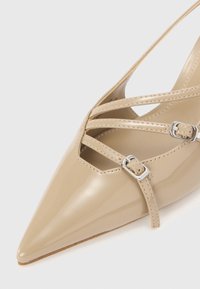 Scarpe in vernice beige con punta a punta e cinturini incrociati con fibbie argentate. La superficie liscia e il design elegante aumentano il fascino moderno.
