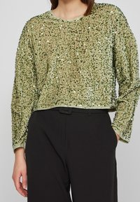 Zielony pulower ozdobiony cekinami, z teksturowanym wykończeniem, okrągłym dekoltem i obniżonymi ramionami. W połączeniu z czarnymi spodniami.