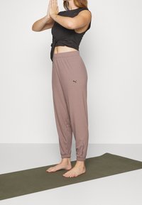 Hellbraune Ripp-Hosen mit elastischem Bund, ausgestattet mit dem schwarzen Puma-Logo, kombiniert mit einem schwarzen Cropped-Top. Das Model befindet sich in einer Yoga-Positur.
