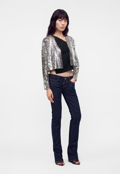 Femme portant une veste courte argentée à sequins sur un haut noir, un jean bleu foncé et des talons noirs ouverts, debout devant un fond uni.