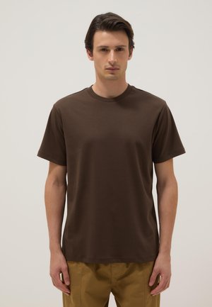 Uomo che indossa una semplice t-shirt marrone scuro a maniche corte e pantaloni beige, in piedi davanti a uno sfondo chiaro e neutro.