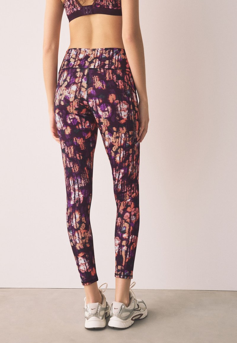 Femme portant des leggings à motifs violets, orange et noirs ainsi qu'un soutien-gorge de sport assorti, debout de dos, avec des chaussures de sport blanches.