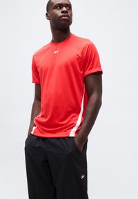 Άνδρας που φοράει κόκκινο αθλητικό κοντομάνικο με λευκό λογότυπο και μαύρο παντελόνι, με τα χέρια στις τσέπες, κοιτάζοντας στο πλάι σε απλό φόντο.