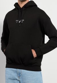 Hombre vistiendo una sudadera con capucha negra con texto blanco y un logo de pájaro en el pecho, con las manos en el bolsillo frontal, combinada con jeans de color azul claro.