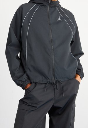 Personne portant une veste noire zippée à capuche avec des bandes réfléchissantes, les mains dans les poches d'un pantalon cargo noir assorti.