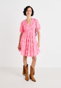 Ροζ φόρεμα με υφασμάτινη φούντα, V λαιμόκοψη, κοντά μανίκια και φαρδύ στυλ. Συνδυασμένο με καφέ καουμπόικες μπότες και τσάντα ώμου με χάντρες.