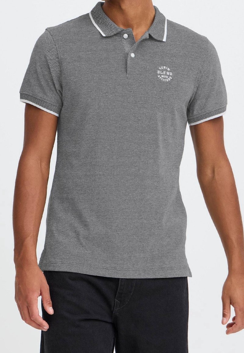 Homme portant un polo gris à manches courtes avec une bordure blanche sur le col et les manches et un petit texte blanc sur la poitrine gauche.