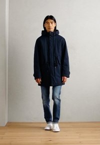 Navy parka with drawstring waist, front pockets, and a hood, paired with light blue jeans and white sneakers on a wooden floor.