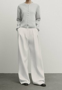 Nainen, jolla on vaaleanharmaa napitettu neuletakki, leveälahkeiset valkoiset housut ja mustat neliövarvaskengät, seisoo käsipuolilla taskuissa.