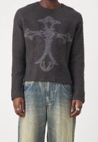 Personne portant un pull gris foncé et duveteux avec un grand motif abstrait en forme de croix et un jean bleu délavé à jambes larges, mains visibles avec des bagues.