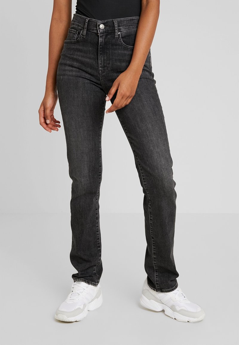 Persona que lleva jeans negros de cintura alta y zapatillas blancas, de pie frente a un fondo gris claro liso.