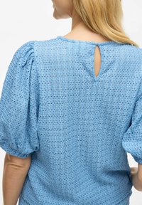 Blusa azzurro chiaro con maniche a palloncino in tessuto texturizzato con piccoli fori, dettaglio a occhiello sulla schiena e vestibilità rilassata.