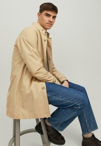 Beige trenchcoat met knoopsluiting, zittend model op een kruk, gekleed in blauwe jeans en donkere schoenen, tegen een lichte achtergrond.