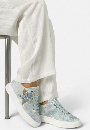 Pies que llevan zapatillas texturizadas azul claro con un detalle de estrella de rhinestones, combinadas con pantalones blancos sueltos, sentado en un taburete de metal.