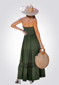 Robe maxi sans bretelles de couleur vert olive avec un corsage smocké, une jupe à volants, des accents en dentelle, et assortie à un sac rond en paille et un chapeau à larges bords.