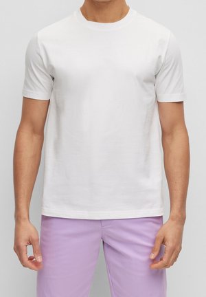 Mann trägt schlichtes weißes kurzärmeliges T-Shirt und helle lila Hose, steht vor hellem grauem Hintergrund.