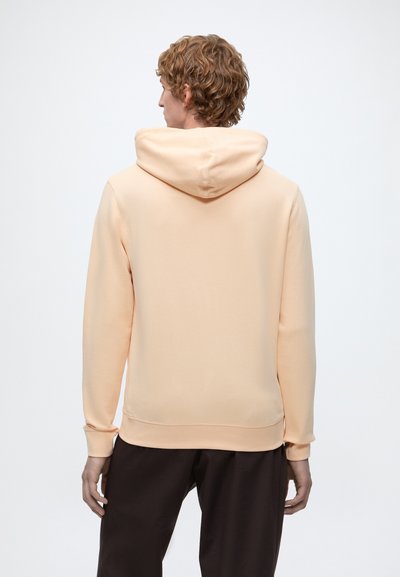 Personne aux cheveux bouclés, vue de dos, portant un sweat à capuche couleur pêche clair et un pantalon foncé sur un fond uni.