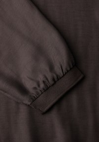 Tissu marron foncé présentant un poignet de manche froncé avec une texture côtelée, une surface lisse et un éclat subtil.