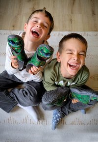 Kinderen houden groene, bedrukte schoenen vast met monsterontwerpen, met grijze zolen en gestructureerde zijkanten, terwijl ze op een gestreepte vloerbedekking zitten.