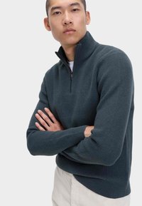 Dunkelblauer, gerippter Strickpullover mit halbem Reißverschluss, langen Ärmeln und taillierter Passform, getragen von einem Modell mit verschränkten Armen.