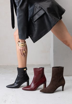 Tři kotníkové boty na podpatku: černá kůže se zlatým zdobením, bordó kůže a hnědá semišová, všechny s špičatou špičkou a různou výškou podpatku.