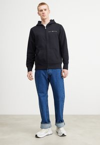 Felpa nera con zip, tasca frontale e cappuccio, con un logo grigio. Abbinata a jeans di denim azzurro chiaro e sneakers bianche.