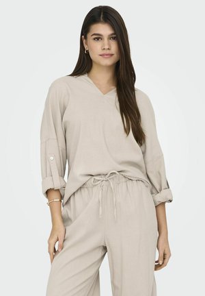 Completo loungewear beige composto da un top a maniche lunghe con scollo a V e polsini arrotolati, abbinato a pantaloni con coulisse, realizzato in tessuto morbido e texturizzato.