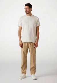 White ribbed short-sleeve t-shirt paired with beige trousers and white sneakers, standing on a light background, arms relaxed by sides.
