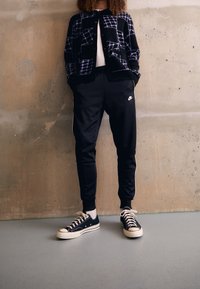 Osoba v černých Nike teplácích, černobílých teniskách a černém vzorovaném kardiganu, stojící u jednoduché betonové zdi.