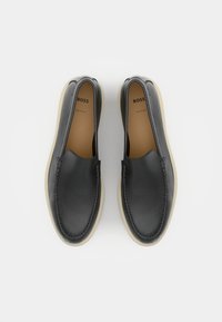 Black leather slip-on shoes with a rounded toe, tan inner lining, and off-white sole. Features subtle stitching and minimalist design.
