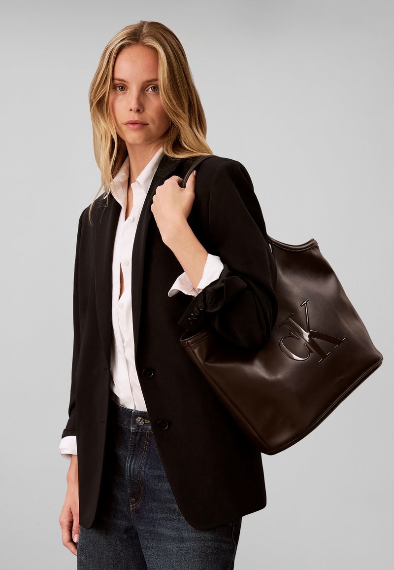 Femme portant un blazer noir et une chemise blanche tenant un grand sac fourre-tout en cuir marron foncé avec un logo CK embossé sur l'épaule.